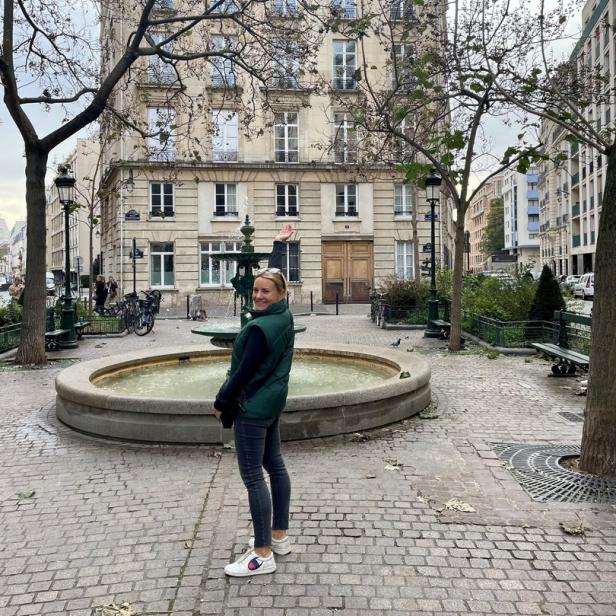 Eine Frau steht auf einem Platz in Paris mit einem Brunnen im Hintergrund.