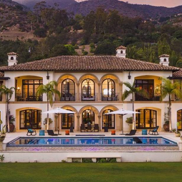 Ein großes, luxuriöses Haus mit Pool vor einer Hügellandschaft bei Sonnenuntergang.