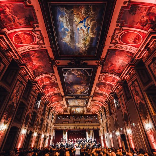 Ein Orchester spielt vor Publikum in einem prunkvollen Saal mit Deckenmalereien.