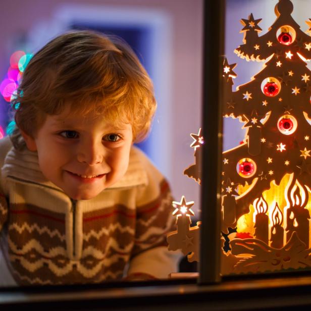 Ein lächelnder Junge schaut durch das Fenster auf einen hölzernen Weihnachtsbaum.