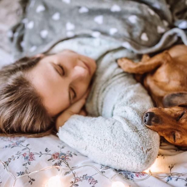 Eine Frau schläft mit ihrem Dackel im Bett, das mit Lichterketten dekoriert ist.