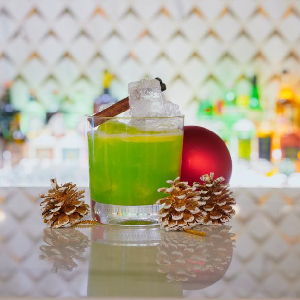 Ein grüner Cocktail mit Eis, Zimtstange und Tannenzapfen vor einer roten Weihnachtskugel.