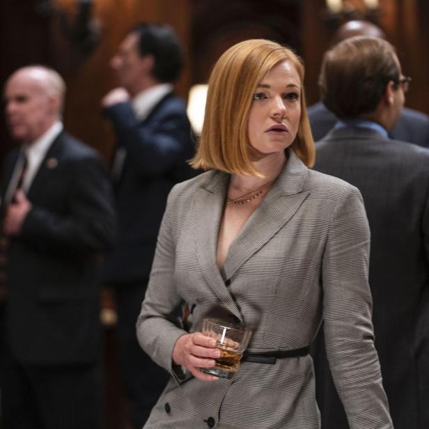 Shiv Roy, gespielt von Sarah Snook, hält ein Glas Whiskey in einer formellen Umgebung.