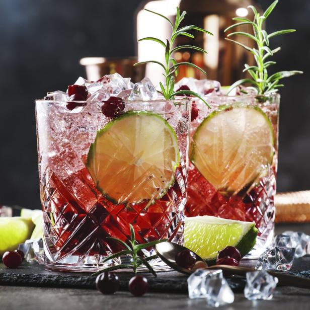 Zwei Gläser mit einem roten Cocktail, Limetten und Eiswürfeln auf einem dunklen Tisch.