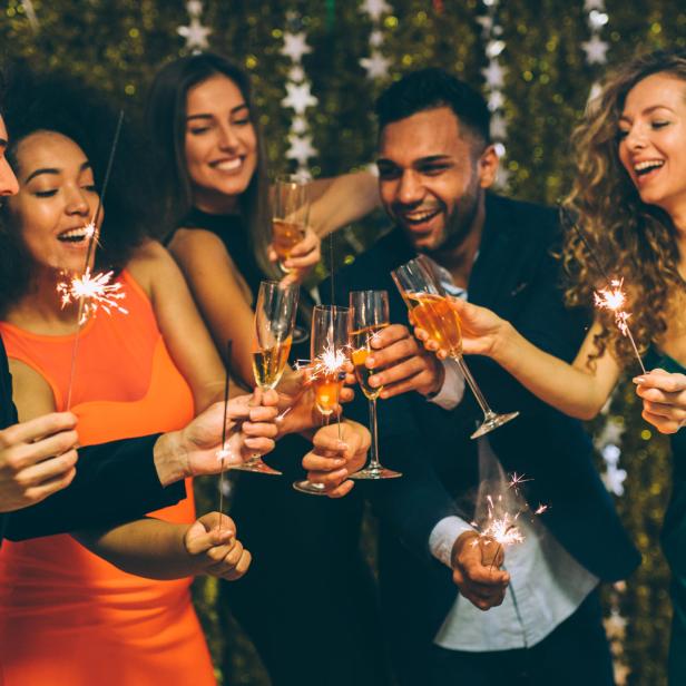 Eine Gruppe junger Leute feiert mit Wunderkerzen und Sekt vor einem goldenen Hintergrund.