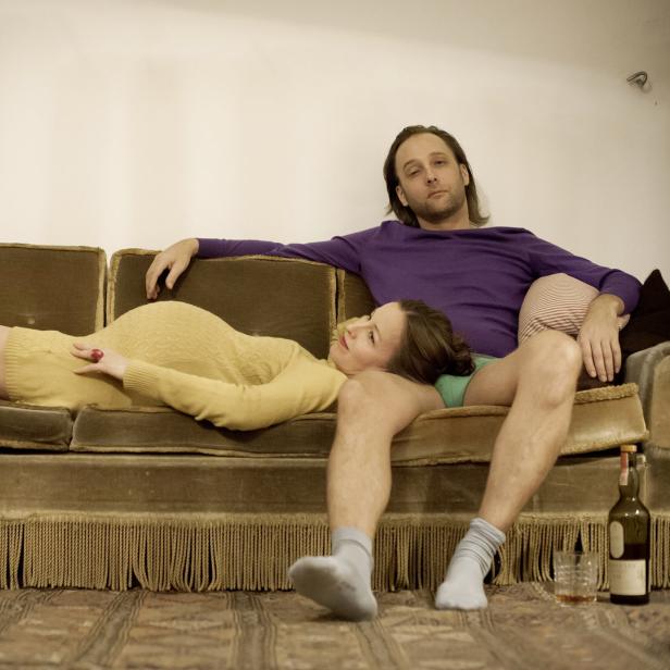 Eine schwangere Frau liegt auf einem Sofa, während ein Mann daneben sitzt.