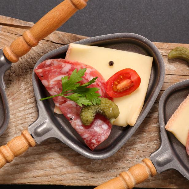 Raclette-Pfännchen mit Käse, Salami, Tomaten und Gewürzgurken auf einem Holztisch.