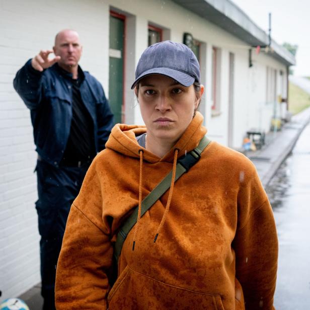 Eine Frau in einem orangefarbenen Hoodie steht im Regen, während ein Mann im Hintergrund auf sie zeigt.