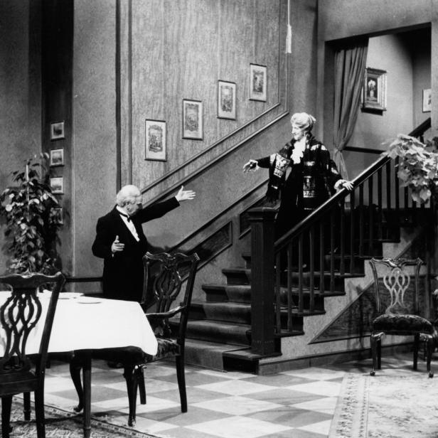 Ein Butler begrüßt eine Dame, die eine Treppe in einem eleganten Haus herunterkommt.