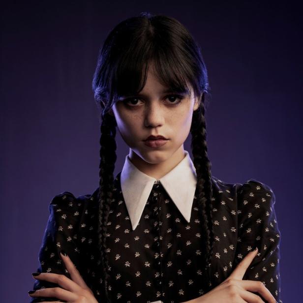 Jenna Ortega als Wednesday Addams mit verschränkten Armen vor einem violetten Hintergrund.