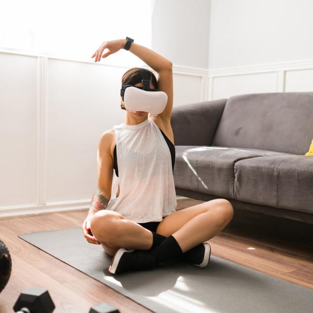 Eine Frau sitzt mit einer VR-Brille auf einer Yogamatte und dehnt sich.