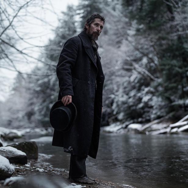 Christian Bale steht in einem langen Mantel an einem Fluss im Winter.
