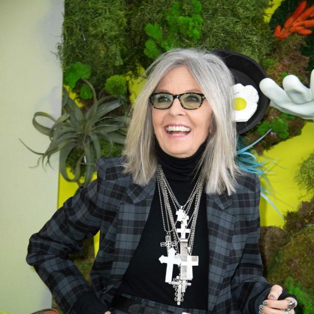 Diane Keaton in einem karierten Hosenanzug und mehreren Kreuzketten lächelt.