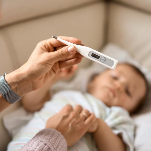 Worried young mother sitting on sofa beside her sick son with high fever. Mom measures temperature using thermometer of sick child lying under blanket at home