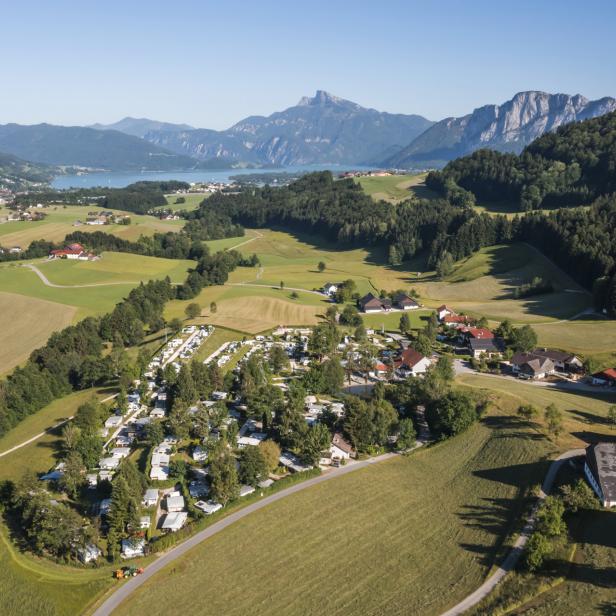 Luftaufnahme eines Campingplatzes in einer grünen Landschaft mit Bergen und einem See im Hintergrund.