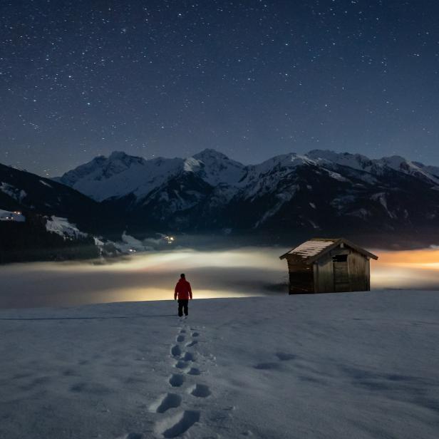 Eine Person geht im Schnee auf eine Hütte zu, im Hintergrund die Berge unter dem Sternenhimmel.