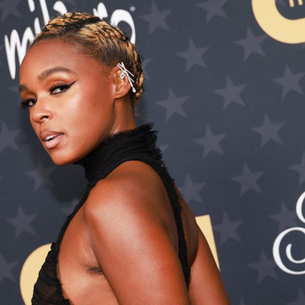 Janelle Monáe posiert auf einem Event mit einem schwarzen Kleid und blonden Zöpfen.