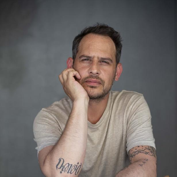 Ein Mann mit Tattoos sitzt auf einem Stuhl und stützt seinen Kopf in die Hand.