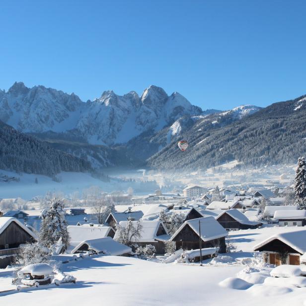 Winterliche Landschaft mit schneebedeckten Häusern und Bergen im Hintergrund. Ein Heißluftballon schwebt in der Ferne.