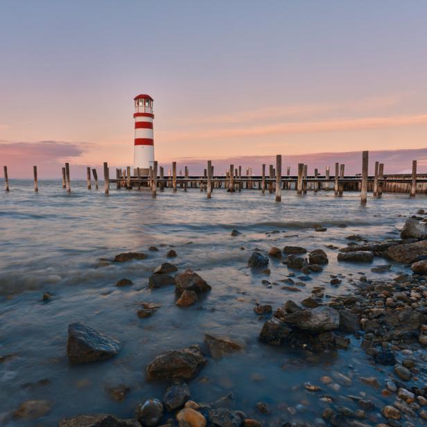 Ein rot-weiß gestreifter Leuchtturm steht an einem See mit einer alten Seebrücke im Hintergrund.