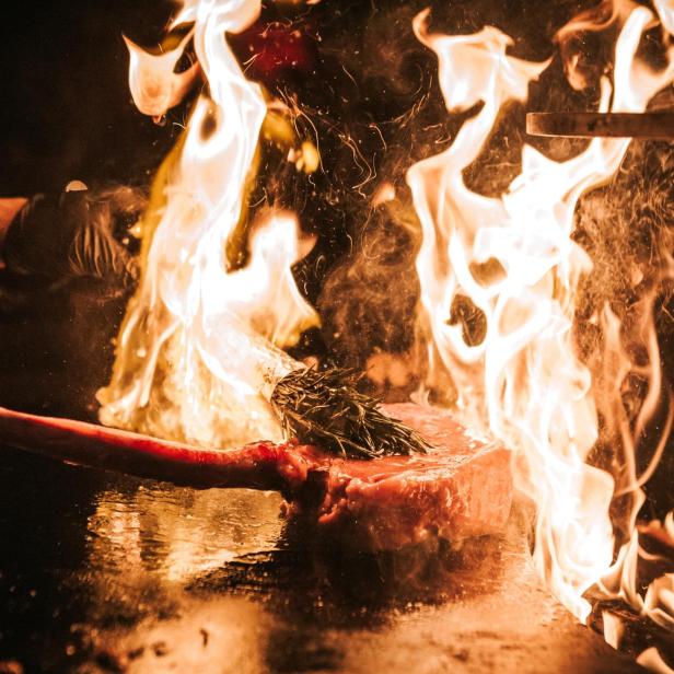 Ein Koch flambiert ein großes Steak mit Rosmarin auf einer heißen Platte.