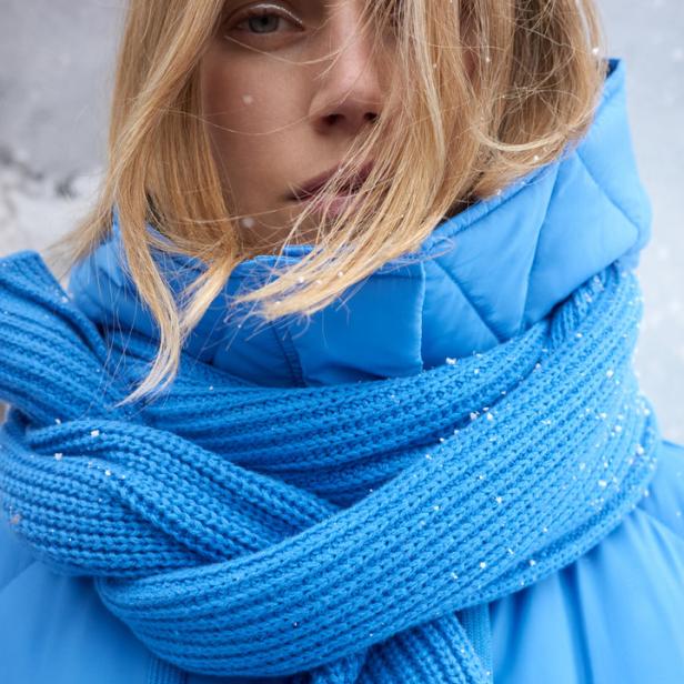 Eine Frau mit blonden Haaren trägt einen blauen Schal und eine passende Jacke im Schnee.
