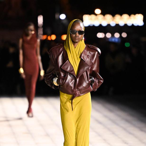 Eine Frau mit Sonnenbrille und Kopftuch trägt eine Lederjacke über einem gelben Kleid auf dem Laufsteg.