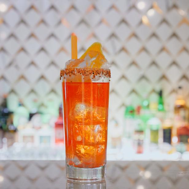 Ein orangefarbener Cocktail mit Eiswürfeln, Strohhalm und einer Orangenscheibe an einer Bar.
