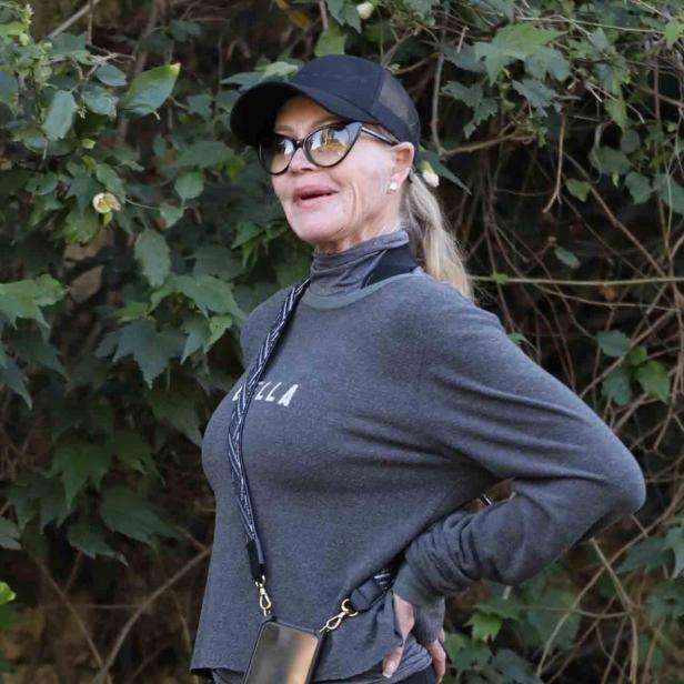 Eine Frau mit Kappe, Brille und Handy-Umhängetasche steht vor einer grünen Hecke.