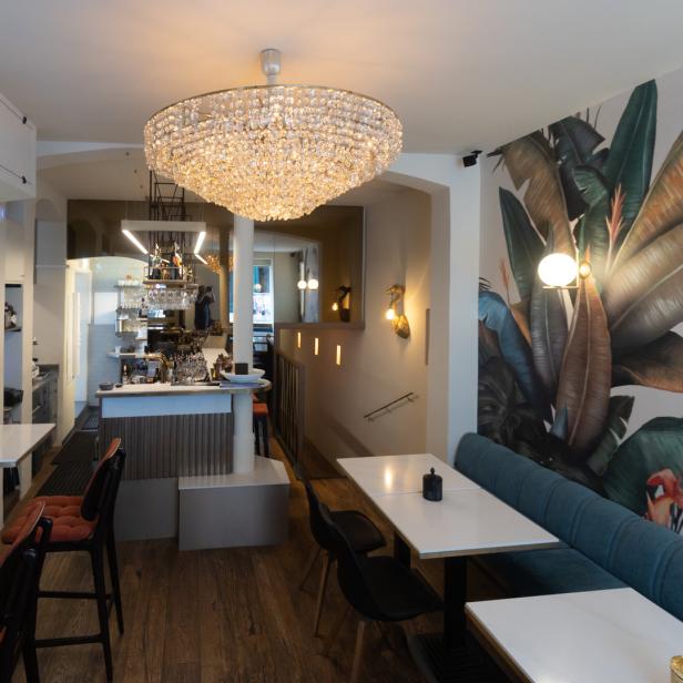Innenansicht eines Restaurants mit Tischen, Stühlen, einer Bar und einer großen Kristalllampe.