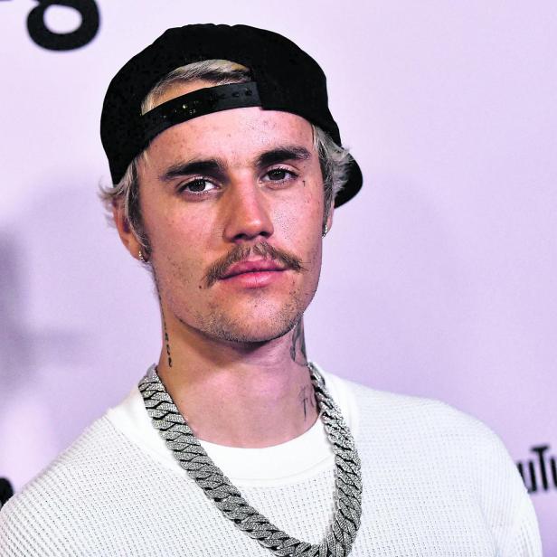 Justin Bieber mit Kappe und auffälliger Halskette vor einem lila Hintergrund.