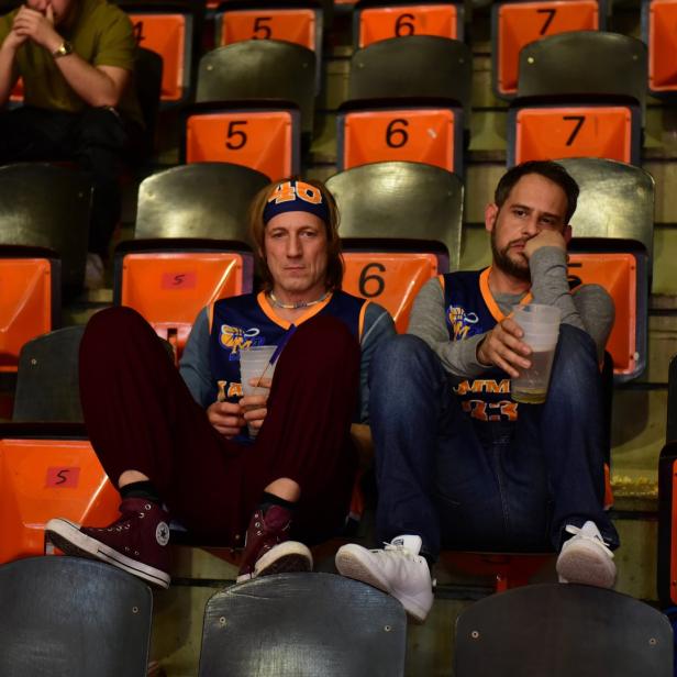 Zwei gelangweilte Männer sitzen mit Getränken auf nummerierten Sitzen in einem Stadion.