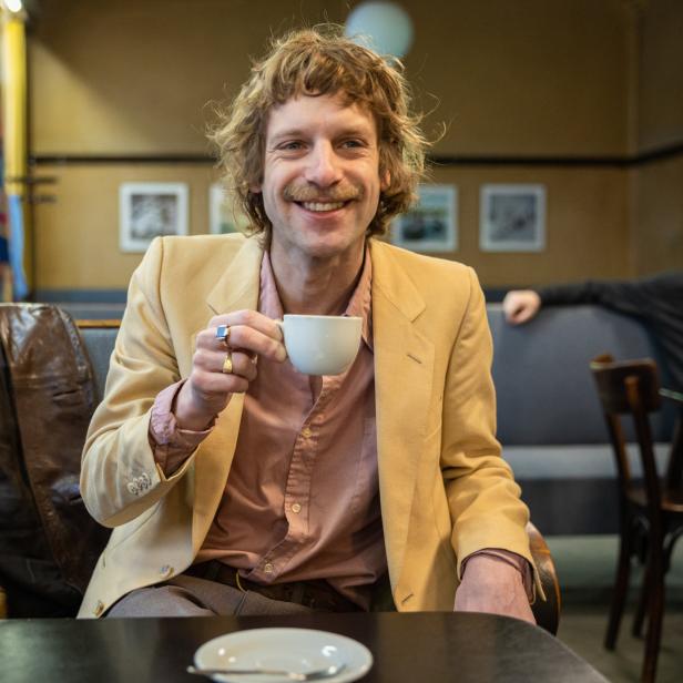 Ein lächelnder Mann mit Schnurrbart sitzt in einem Café und hält eine Tasse in der Hand.
