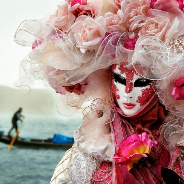 Eine Person in Venedig trägt eine aufwendige Karnevalsmaske und einen Rosenkopfschmuck. Im Hintergrund eine Gondel.