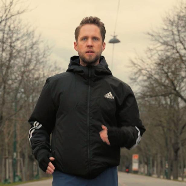 Ein Mann mit Adidas-Jacke joggt auf einer von Bäumen gesäumten Straße.