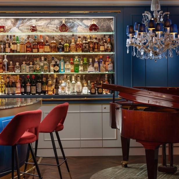 Eine Bar mit Spirituosenregal, roten Barhockern und einem Klavier unter einem Kronleuchter.