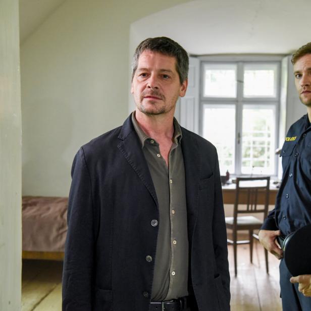 Ein Mann in Zivil steht neben einem österreichischen Polizisten in einem Haus.