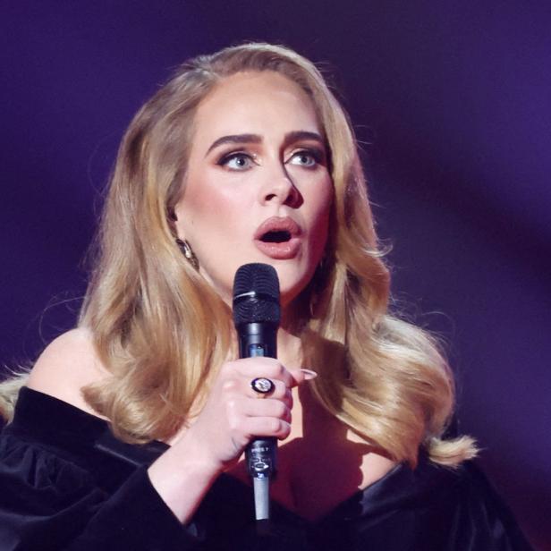 Adele singt mit einem Mikrofon in der Hand vor einem violetten Hintergrund.