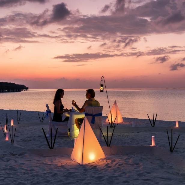Ein Paar genießt ein romantisches Abendessen am Strand bei Sonnenuntergang, beleuchtet von Laternen.