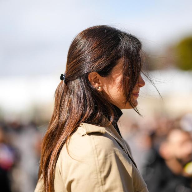 Eine Frau mit braunen Haaren, die zu einem Pferdeschwanz zusammengebunden sind, trägt einen Trenchcoat.