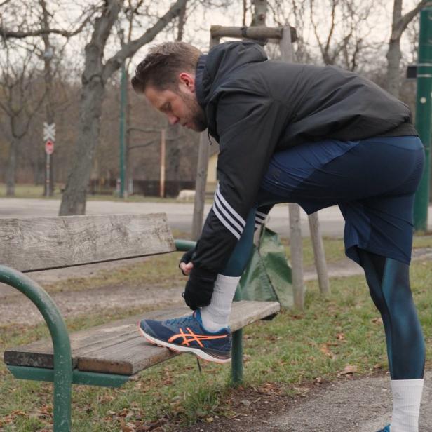 Ein Mann bindet sich auf einer Parkbank die Schnürsenkel seiner Laufschuhe.