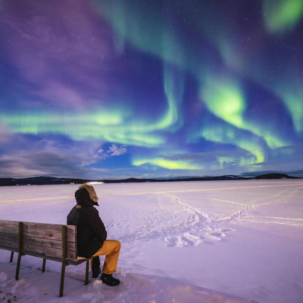 Eine Person sitzt auf einer Bank und beobachtet die Nordlichter über einer verschneiten Landschaft.
