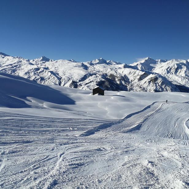 Eine kleine Hütte steht auf einer schneebedeckten Berglandschaft unter blauem Himmel.