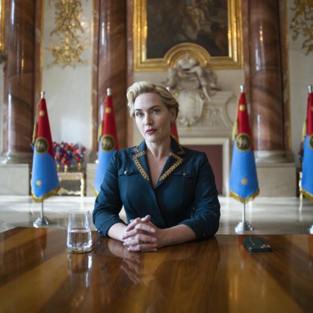 Kate Winslet sitzt an einem Tisch vor Flaggen in einem opulenten Raum.