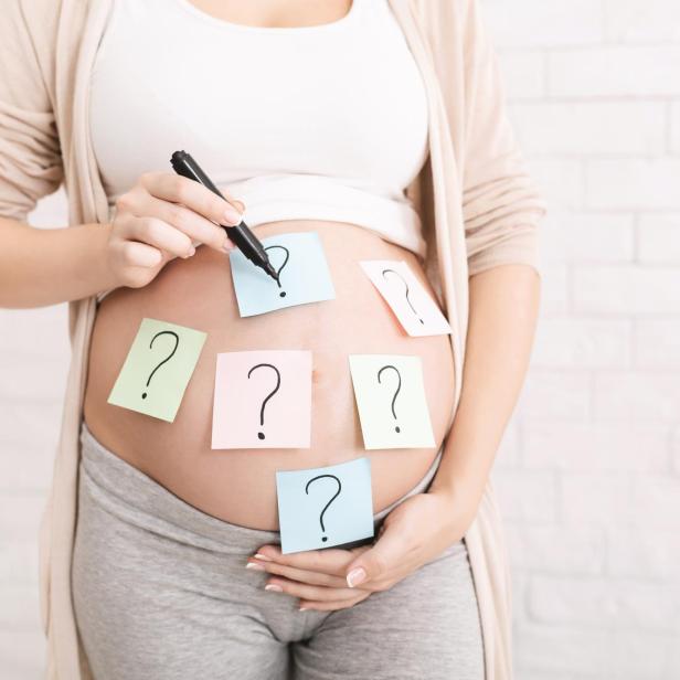 Eine schwangere Frau mit Fragezeichen-Notizen auf ihrem Bauch hält einen Stift.