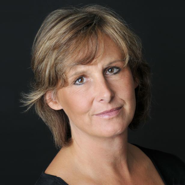 Porträt einer Frau mit blonden, gewellten Haaren vor einem dunklen Hintergrund.