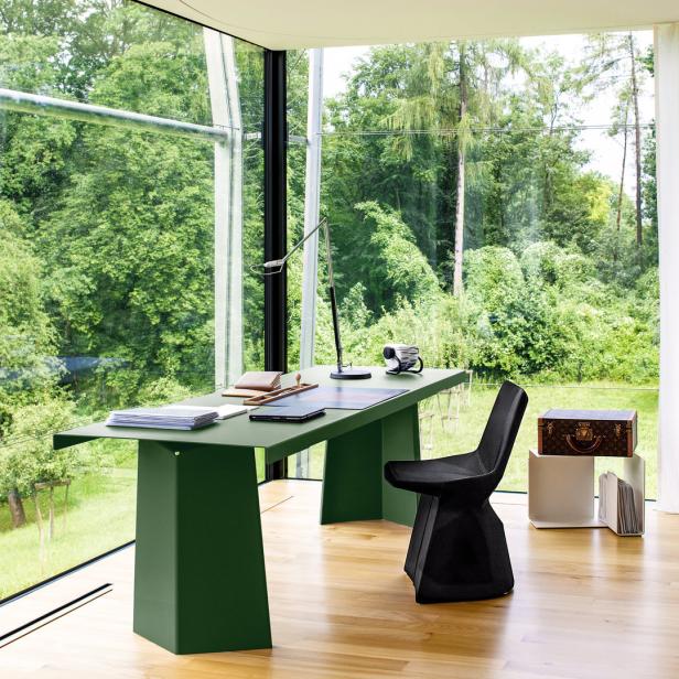 Ein grüner Schreibtisch mit Büromaterialien steht vor einem großen Fenster mit Blick auf einen grünen Garten.
