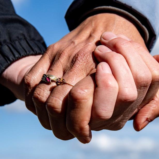 Zwei Menschen halten Händchen vor einem Himmel mit Wolken. Eine Hand trägt einen goldenen Ring mit einem roten Stein.