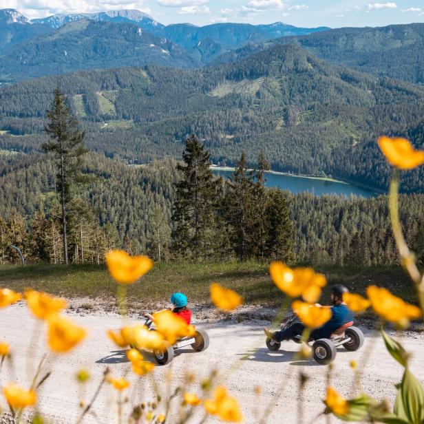 Zwei Personen fahren mit Mountaincarts auf einer Schotterstraße vor einer Bergkulisse.