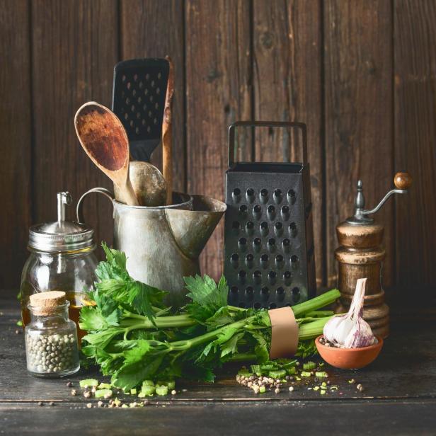 Rustikales Kochset mit Vintage-Küchenutensilien und Selleriebündel auf dunklem Tisch vor Holzgrund.